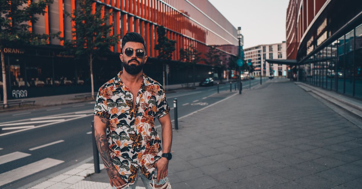 Homme stylé avec des tatouages et des lunettes de soleil posant avec confiance dans un cadre urbain moderne.