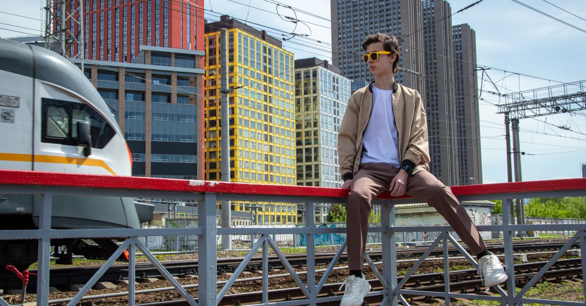 Un jeune homme à la mode avec des lunettes de soleil est assis près des voies ferrées avec une vue sur la skyline de la ville.