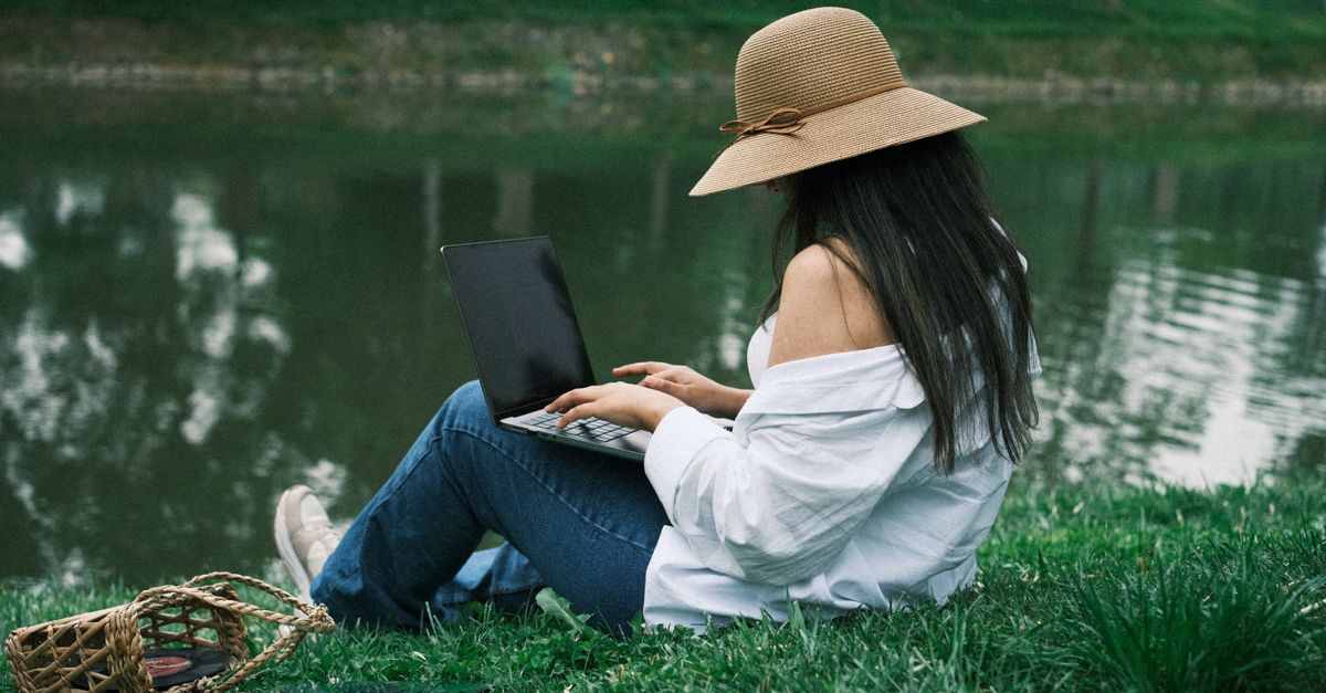 Une femme est assise près d'un lac, travaillant sur son ordinateur portable dans un parc paisible.
