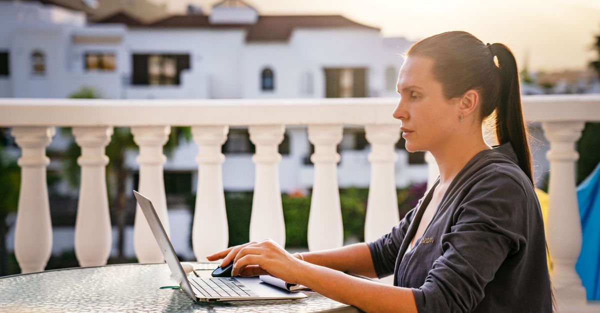 Une femme concentrée travaille sur son ordinateur portable à l'extérieur sur un balcon ensoleillé, capturant une ambiance de travail sereine du matin.