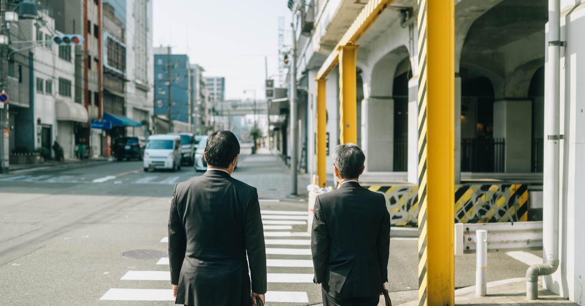 Deux hommes d'affaires en costume traversant une rue dans un décor urbain japonais, reflétant la vie quotidienne de la ville.