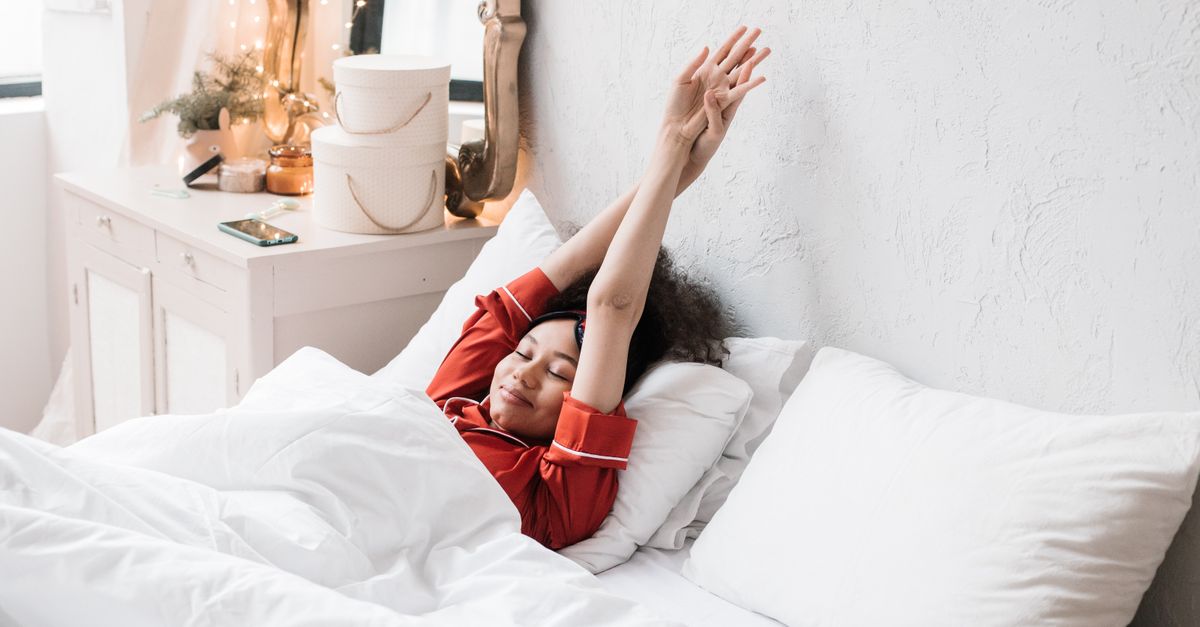 Femme en pyjama rouge s'étirant dans une chambre minimaliste et confortable. La lumière matinale éclatante met en valeur la scène sereine.
