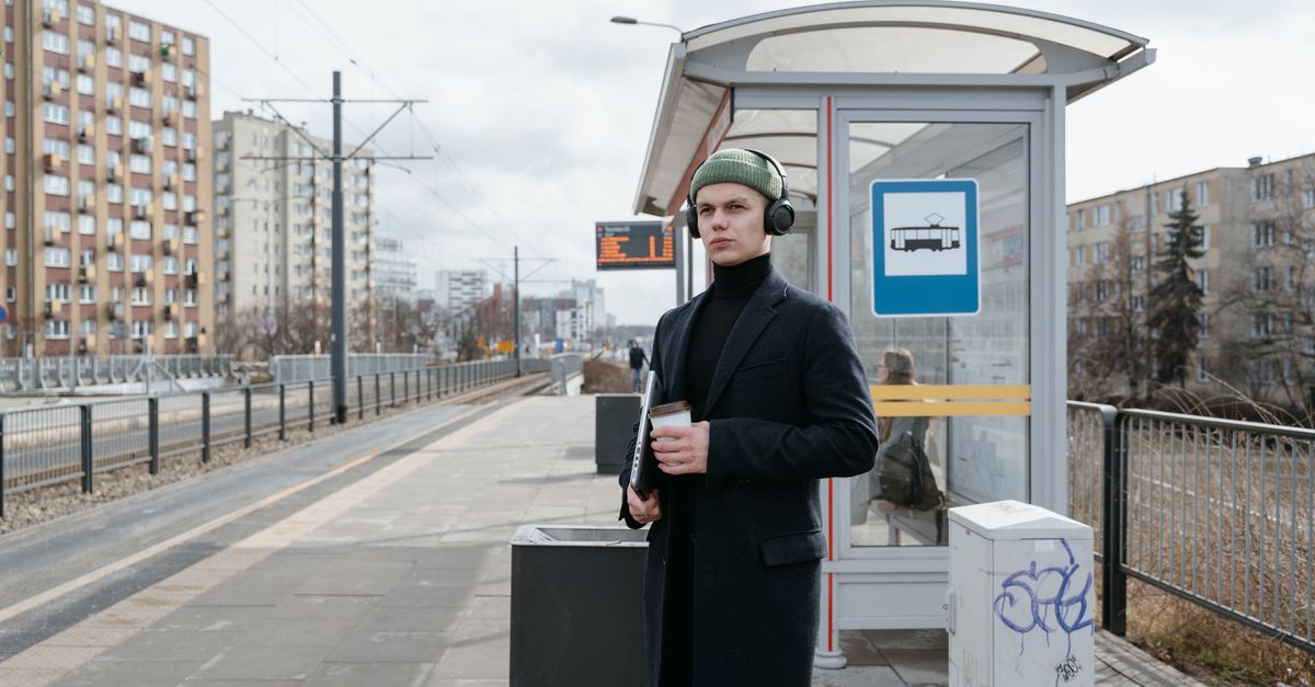 Jeune homme en manteau et écouteurs tenant un café à la gare extérieure, en journée.