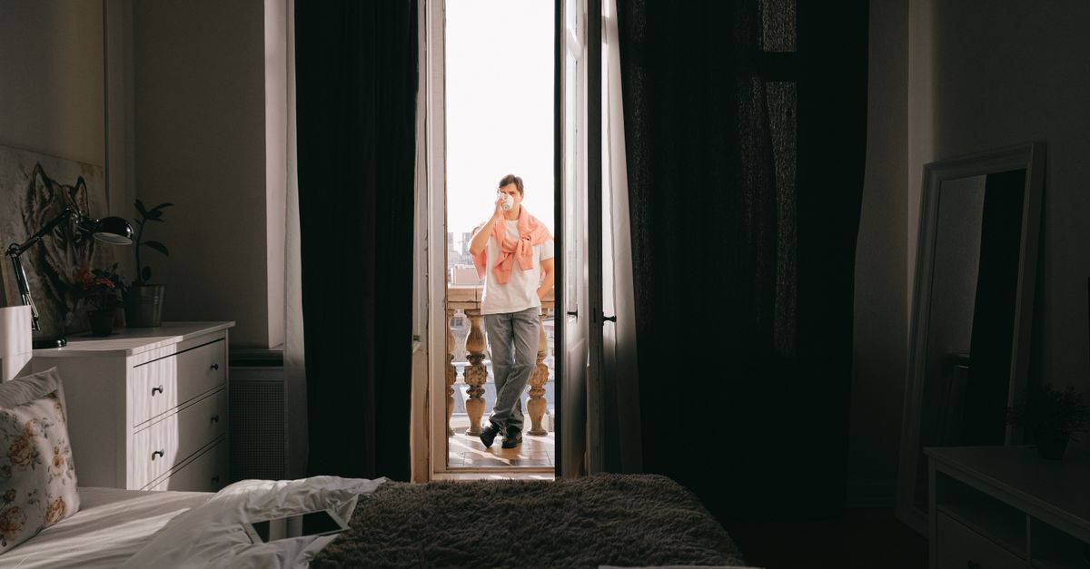 Un homme se tient sur un balcon dégustant un café, la lumière du matin pénétrant dans une chambre cozy.