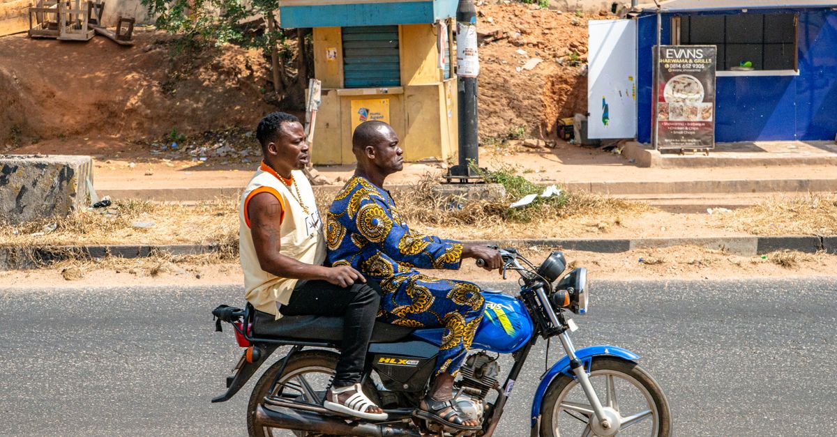 Deux hommes sur une moto parcourent une rue urbaine ensoleillée, mettant en valeur la vie quotidienne et les transports.